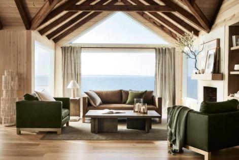 Bright living room with wooden beams, green armchairs, and ocean views through large windows.