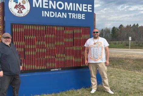 Two people stand smiling beside a large blue sign that reads “Menominee Indian Tribe,” with trees and a road visible in the background.