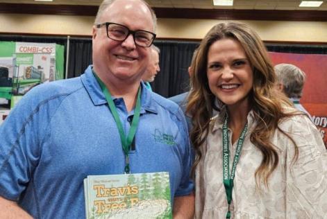 Two people smile at an event, one holding a children’s book titled Travis Visits the Tree Farm.