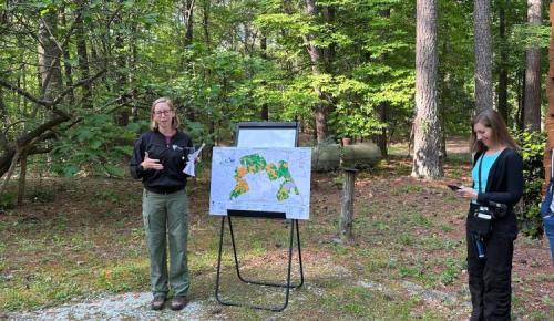 A speaker gestures beside a large map on an easel while another person takes notes in a wooded area.