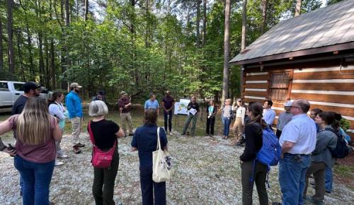 A group of about twenty people stand in a circle outside a log cabin, listening to a speaker during a Duke Forest field event surrounded by tall trees.