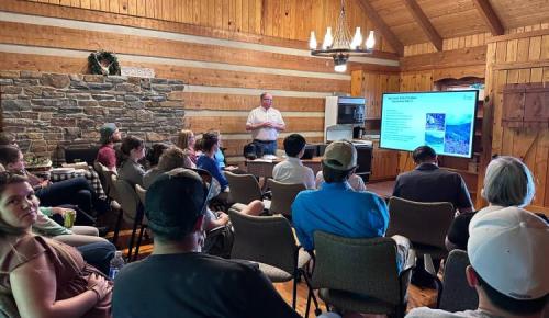 A group sits in a wood-paneled lodge while a speaker presents slides on forest certification.