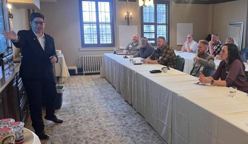 A speaker addresses a group seated in rows, gesturing toward a screen at the front of the room.