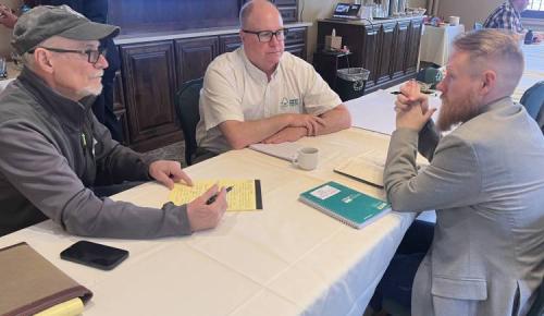 Three men sit at a table with notebooks and coffee, talking during a breakout discussion.
