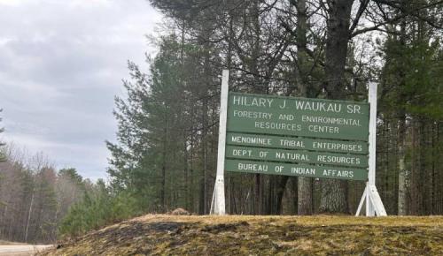 A green roadside sign reads “Hilary J. Waukau Sr. Forestry and Environmental Resources Center” surrounded by pine trees.