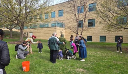 A group of adults and children gather on a grassy lawn beside a small tree, preparing to plant it outside a tan brick building.