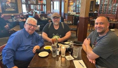 Three people sit at a restaurant table smiling with drinks and plates in front of them.