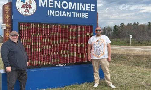 Two people stand smiling beside a large blue sign that reads “Menominee Indian Tribe,” with trees and a road visible in the background.