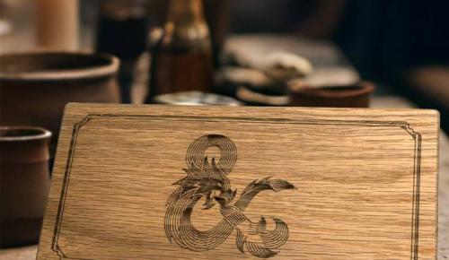 A wooden dice vault with an etched Dungeons & Dragons ampersand dragon logo sits on a tabletop.