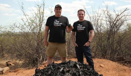 Two people wearing The Good Charcoal shirts stand by a pile of charcoal in a dry landscape.