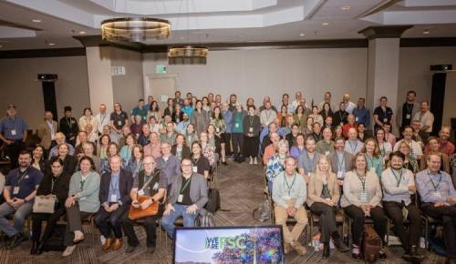 Large group photo of conference participants seated and standing in a meeting room.