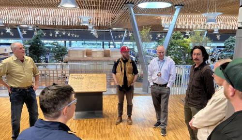 Several people talk near a wood-display table under a curved timber ceiling with trees and architectural models in the background.