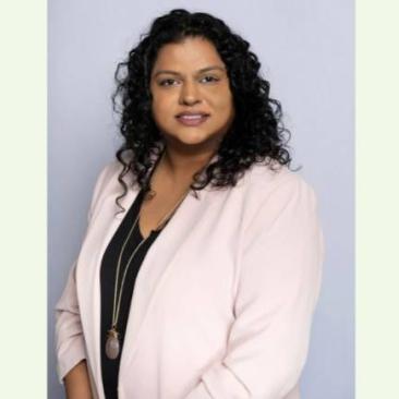 A professional headshot of a woman wearing a light-pink blazer over a black top, smiling against a pale backdrop.